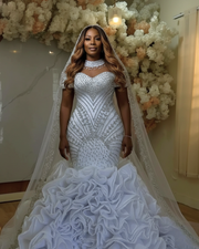Luxurious embellished mermaid wedding dress featuring intricate silver beadwork, illusion neckline with crystal detailing, structured bodice with geometric patterns, detachable embellished cape, and a dramatic multi-layered ruffled skirt in bright white.