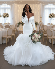 Statement off-the-shoulder mermaid wedding dress featuring a deep plunging neckline, fitted long sleeves, and an extravagant sculpted ruffle skirt with dramatic layered organza detailing in pure white.