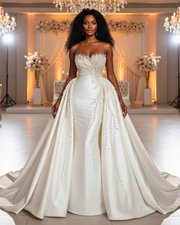 The dress features a fitted silhouette (likely mermaid or trumpet) with an elaborate overskirt. The sweetheart-neckline bodice and the entire front panel of the overskirt are densely hand-embellished with various sizes of clear crystals and white pearls, which appear to cascade downward from the bustline in a dripping effect. The fabric of the skirt and overskirt is a smooth, heavy satin.